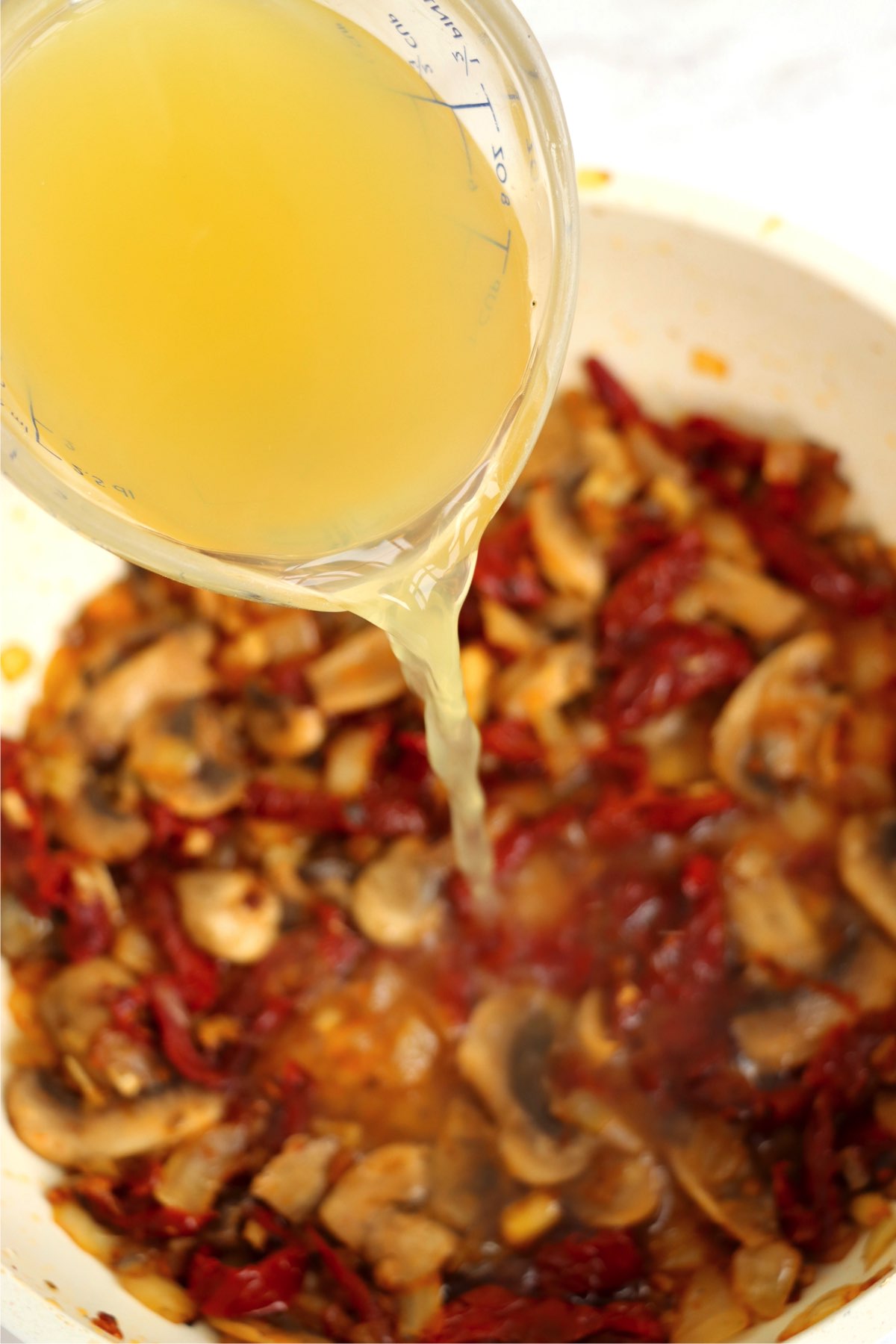 Chicken broth being poured into a skillet filled with the sautéed vegetables and seasonings to create a sauce.