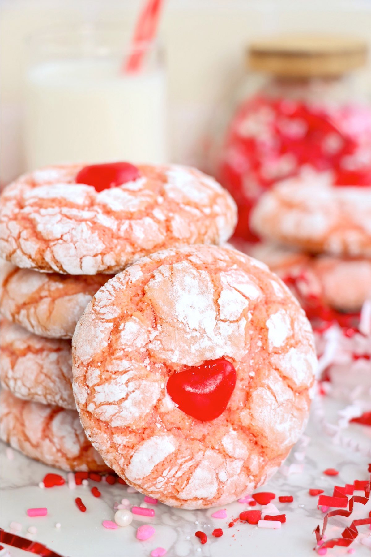 A festive arrangement of strawberry crinkle cookies with a jar of sprinkles and milk in the background.