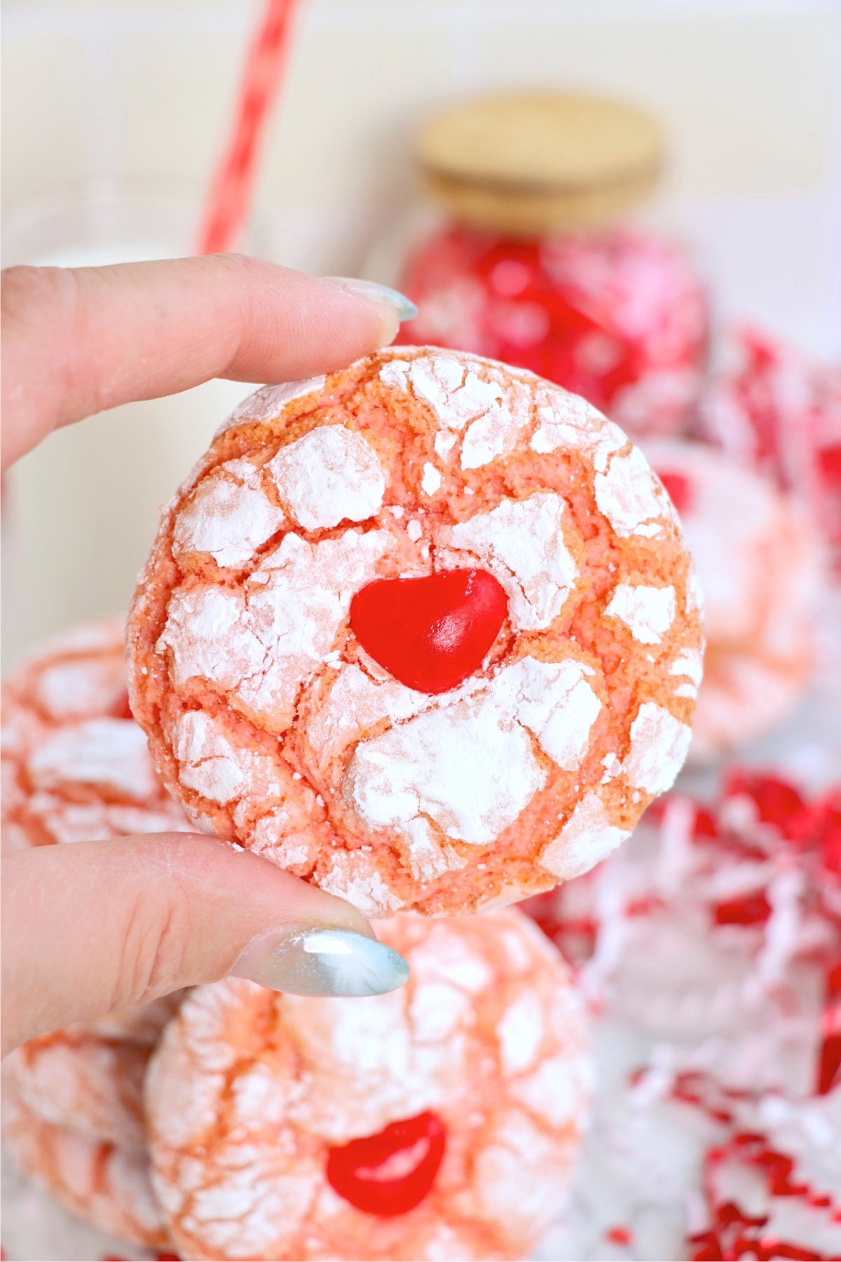 A hand holding a single strawberry crinkle cookie, showcasing the red candy heart and crinkled texture.