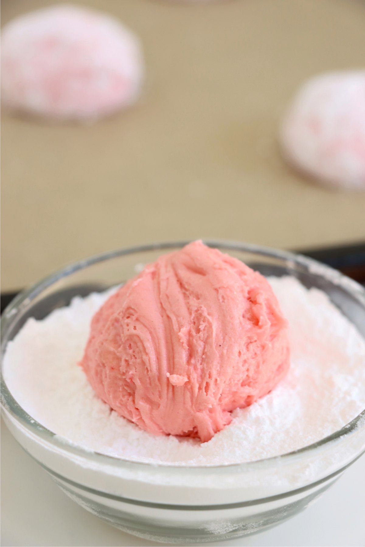 A round ball of strawberry cookie dough being coated in powdered sugar, highlighting the preparation step for achieving the crinkled look.