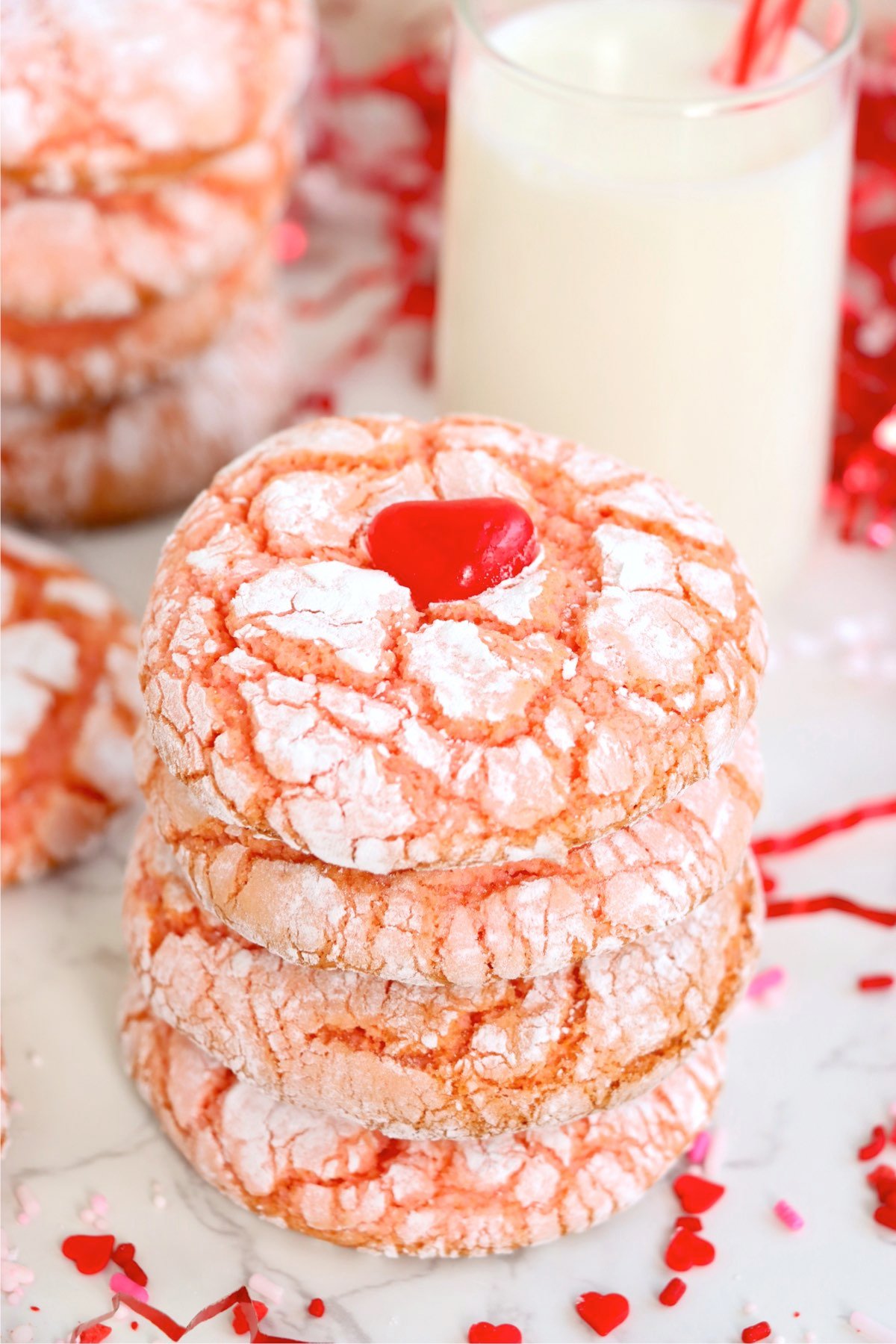 A stack of baked strawberry crinkle cookies topped with a red heart candy, with powdered sugar cracks beautifully visible, surrounded by festive sprinkles and a glass of milk.