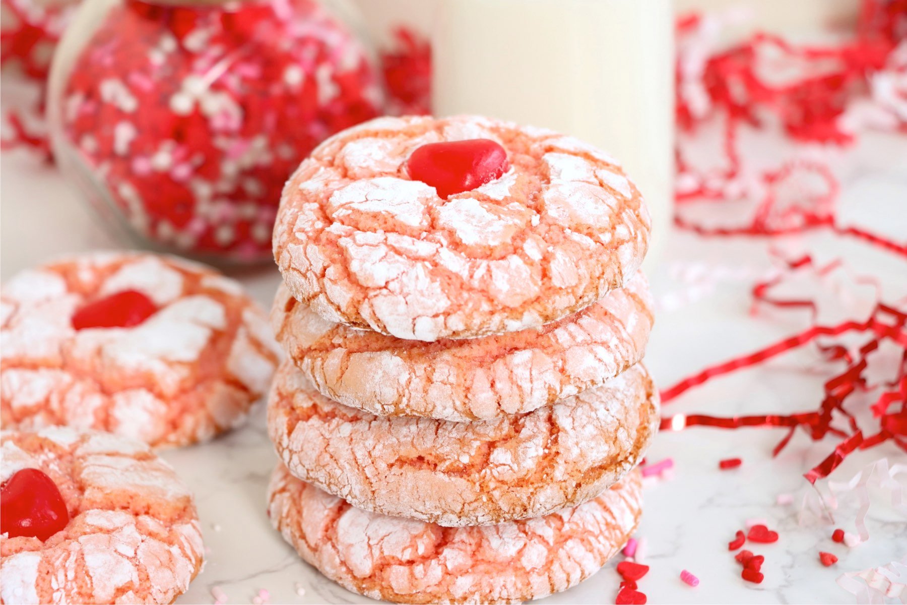 A stack of strawberry crinkle cookies topped with a red candy heart, paired with a glass of milk.