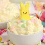 A white bowl of fluffy, creamy dessert with pastel mini marshmallows and a yellow bunny Peep on top, a spoon resting inside the bowl.