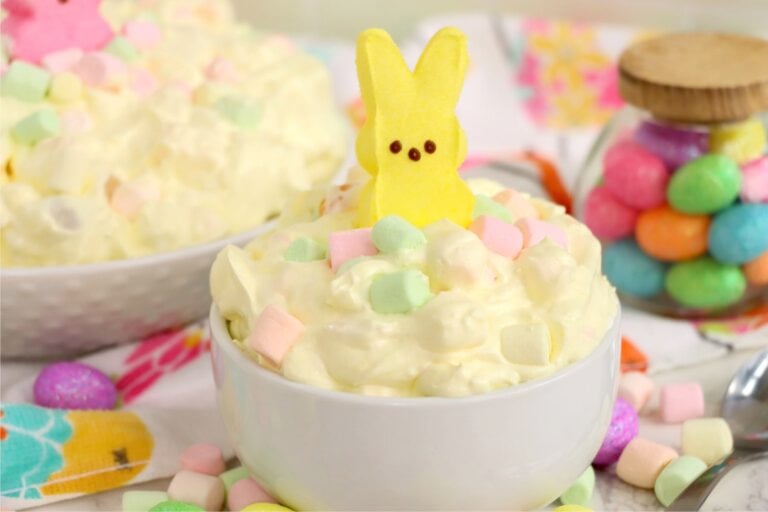 A white bowl of fluffy, creamy dessert with pastel mini marshmallows and a yellow bunny Peep on top, a spoon resting inside the bowl.
