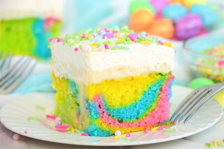 A close-up of a single slice of poke cake with vibrant layers of blue, yellow, green, and pink, topped with a thick layer of whipped cream and sprinkles.