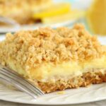 A classic side view of a lemon crumb bar on a white plate, with bright lemon slices and a soft blue background.