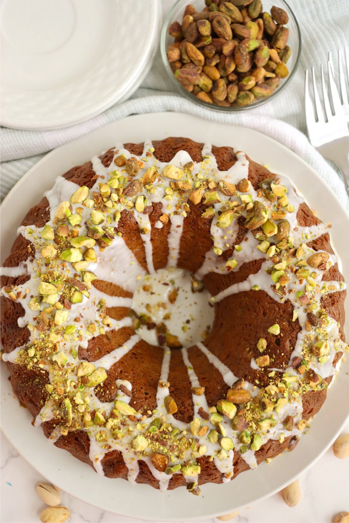 A top-down view of a glazed pistachio bundt cake on a white plate, with a bowl of whole pistachios nearby.