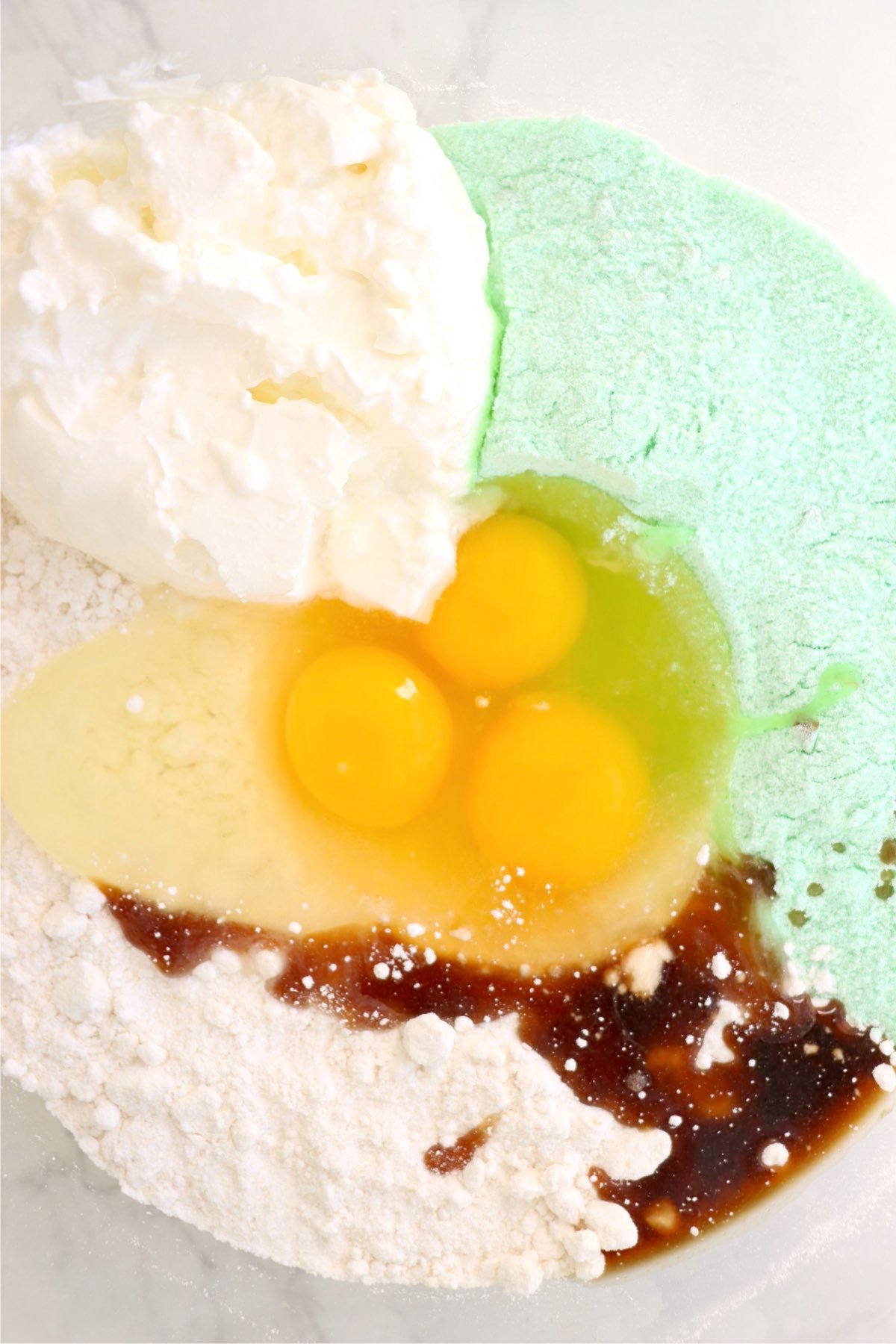 A mixing bowl filled with dry cake mix, pistachio pudding mix, eggs, sour cream, vanilla extract, and vegetable oil, ready to be combined.