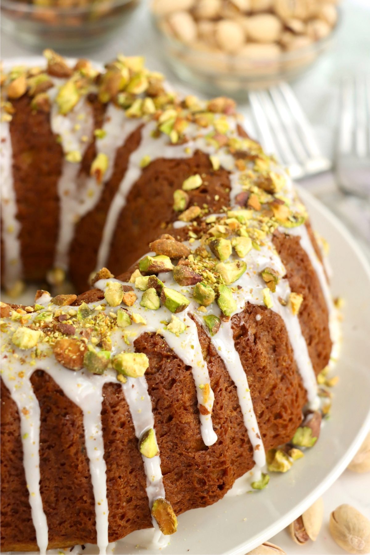 A pistachio bundt cake with a drizzle of icing and crunchy pistachios, with forks and bowls of nuts in the background.