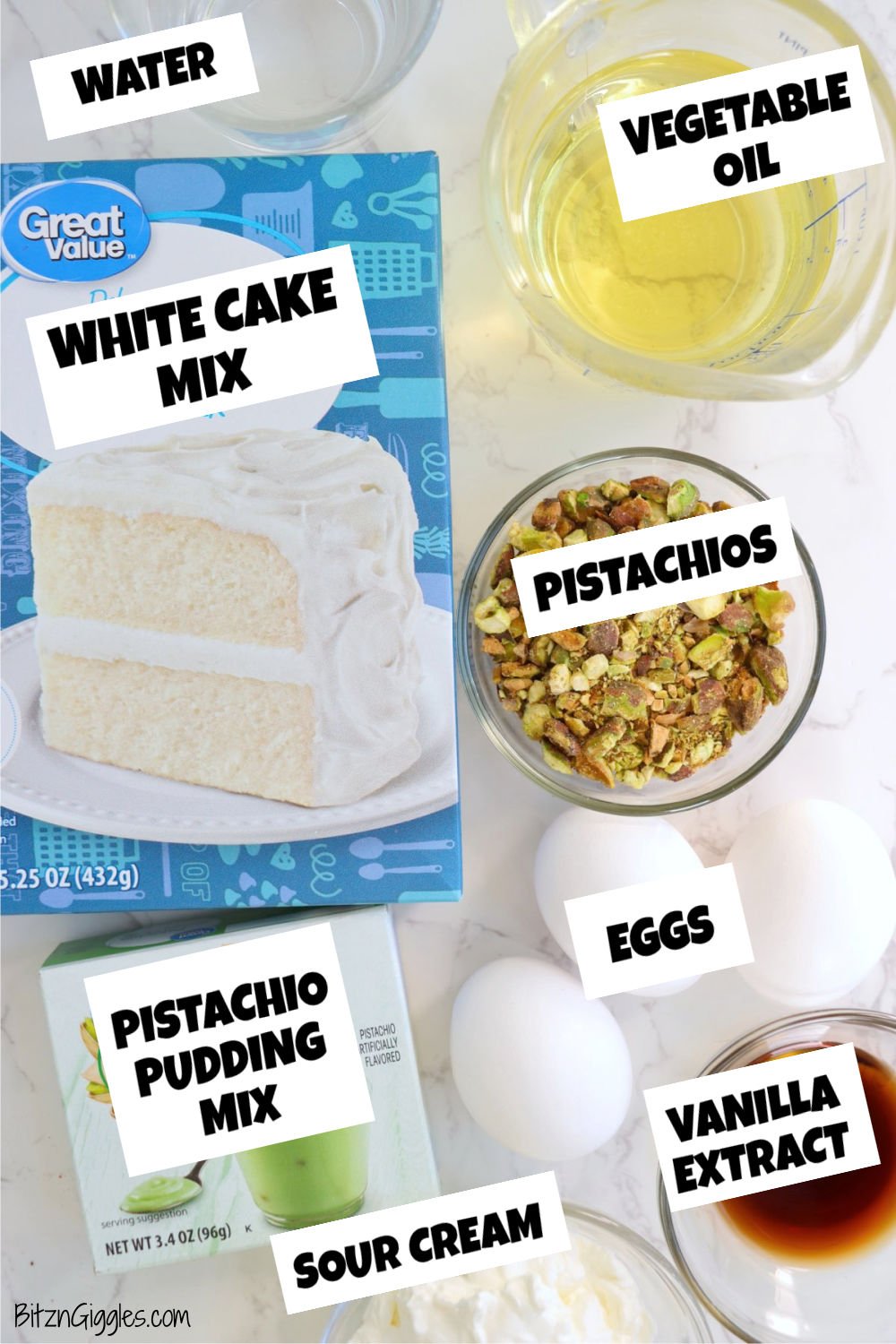 A collection of ingredients laid out on a marble countertop, including a box of white cake mix, a box of pistachio pudding mix, chopped pistachios, eggs, vanilla extract, vegetable oil, sour cream, and water.