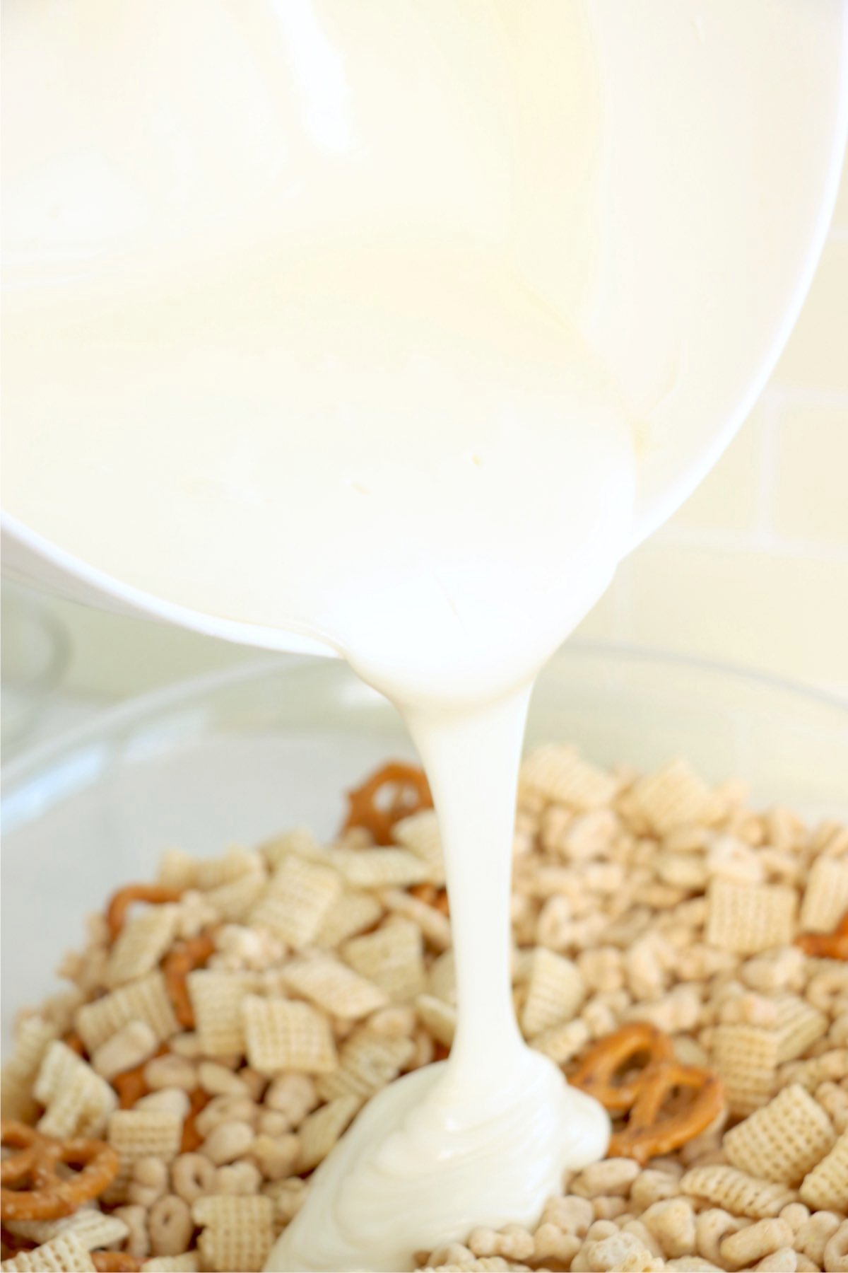 Melted vanilla almond bark being poured over a crunchy mix of Chex cereal, pretzels, and Lucky Charms, creating a sweet and creamy coating.