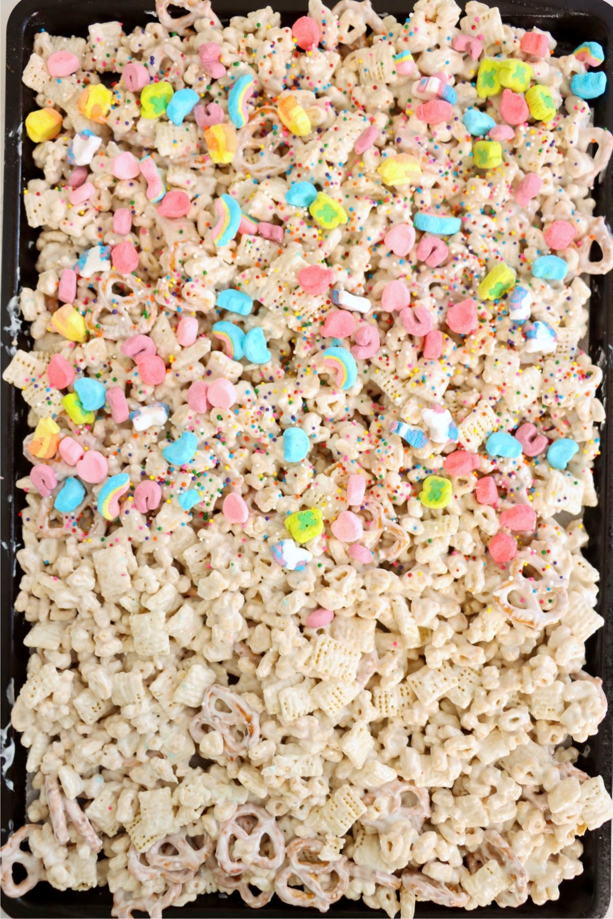 A baking sheet filled with a coated snack mix, topped with colorful Lucky Charms marshmallows and rainbow sprinkles.