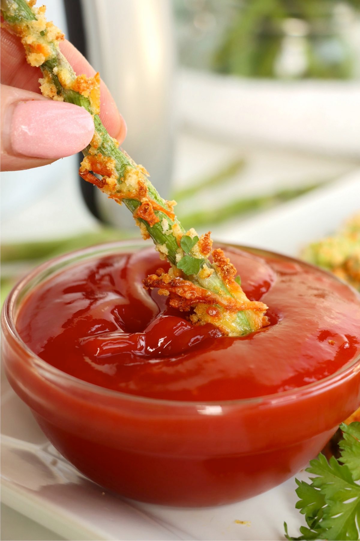 A single crispy asparagus fry being dipped into a bowl of thick ketchup, held between two fingers with a parsley garnish nearby.