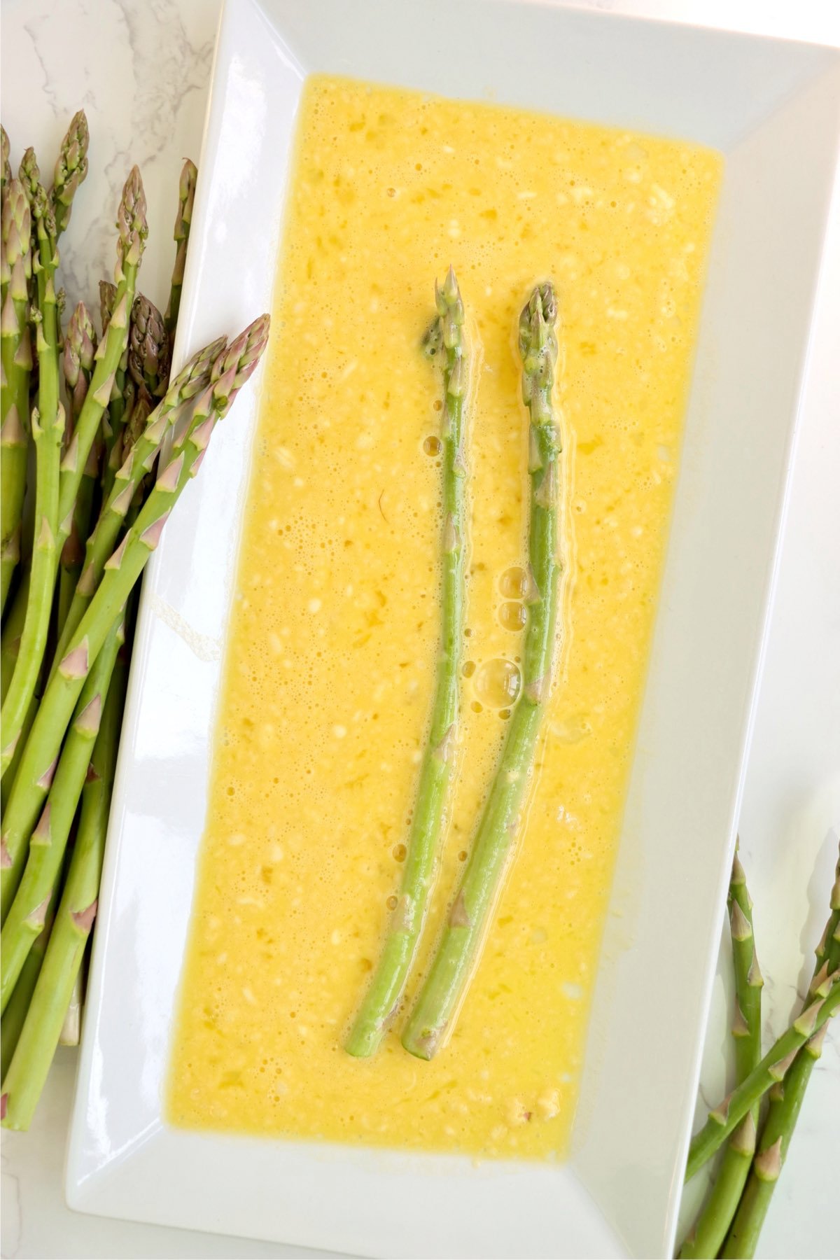 Two asparagus spears dipped in a dish filled with a beaten egg mixture, surrounded by more asparagus.