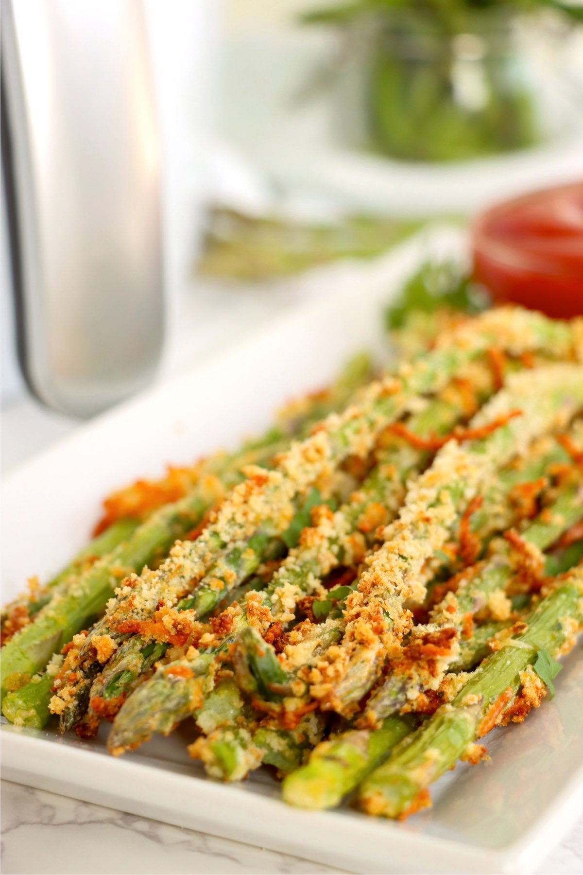 Close-up of crispy asparagus fries on a white tray with a bowl of ketchup and fresh herbs in the background.