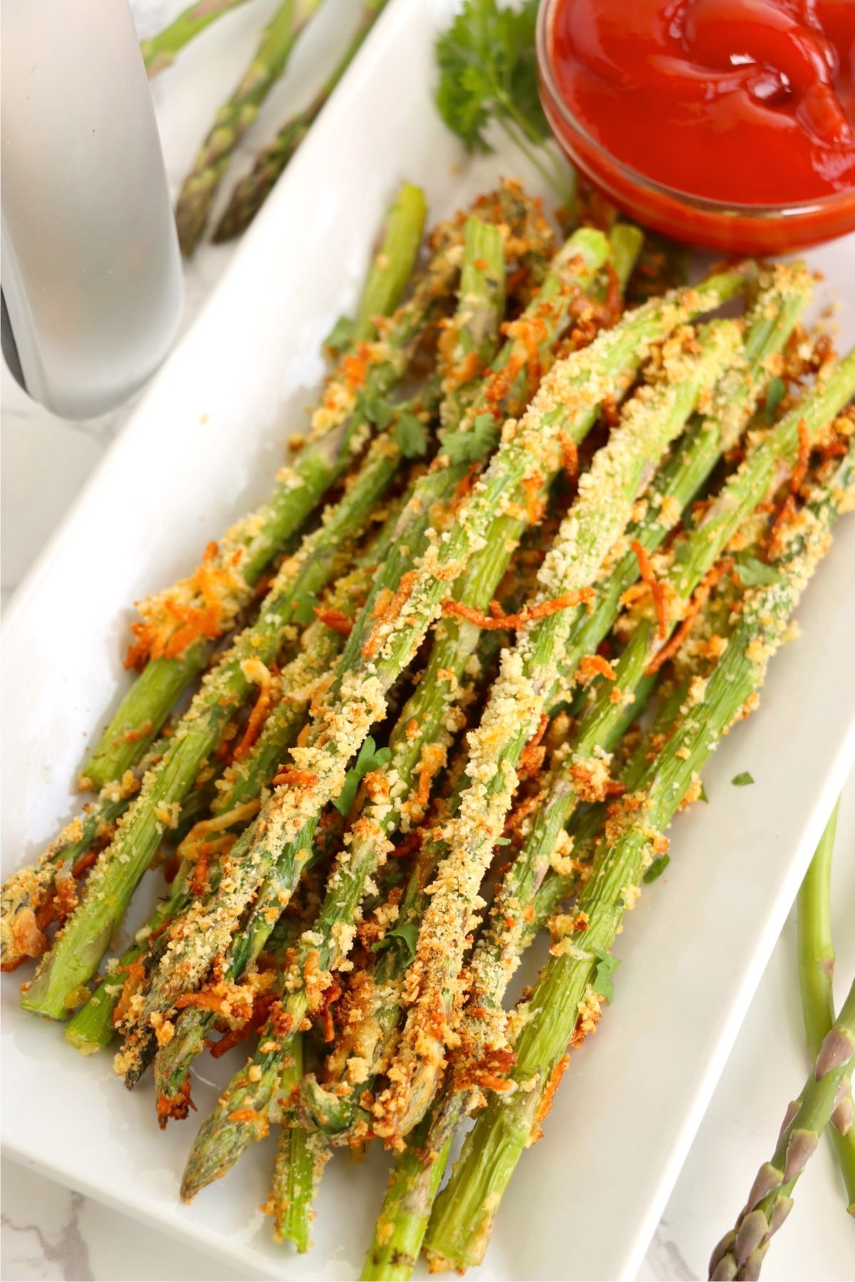 Close-up of crispy asparagus fries on a white tray with a bowl of ketchup and fresh herbs in the background.