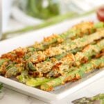 Crispy asparagus fries piled high on a white rectangular plate with parsley and whole asparagus spears nearby.