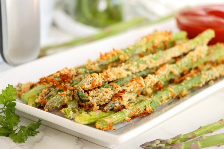 Crispy asparagus fries piled high on a white rectangular plate with parsley and whole asparagus spears nearby.