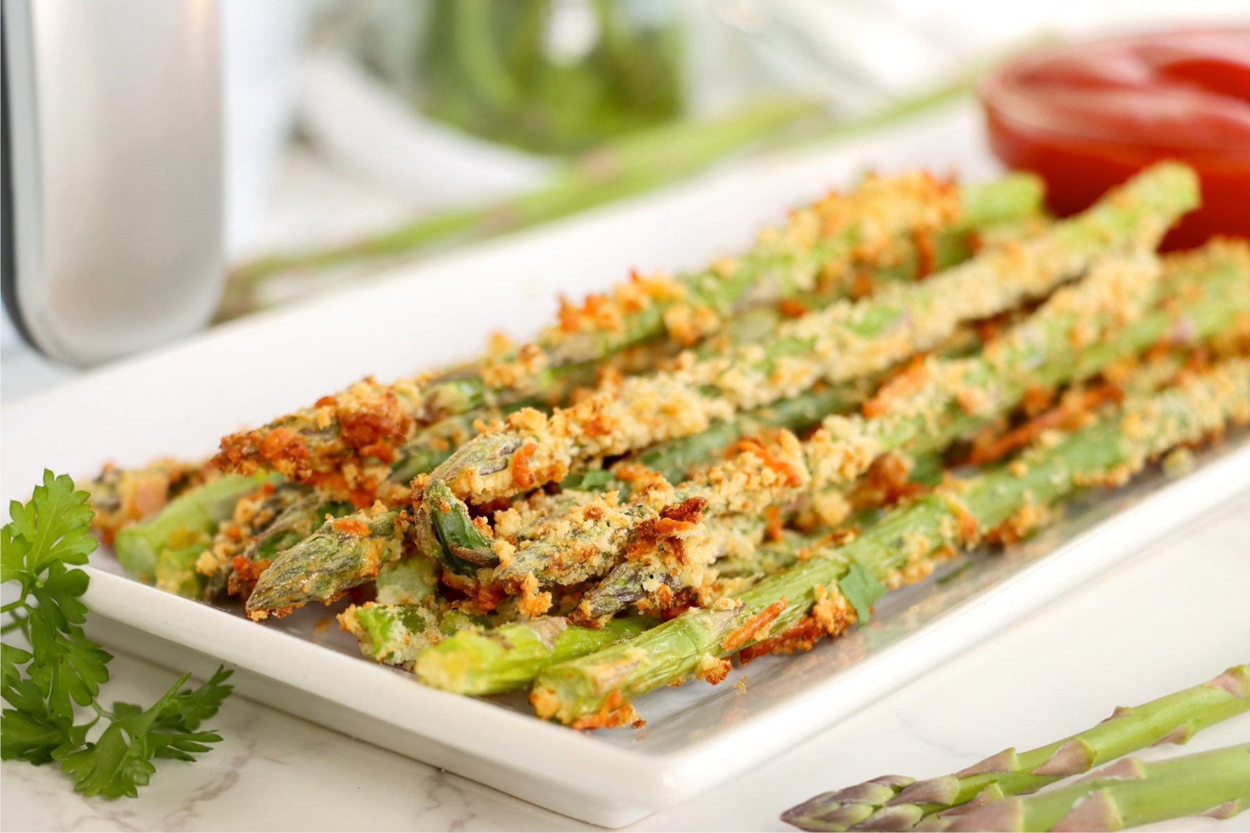 Crispy asparagus fries piled high on a white rectangular plate with parsley and whole asparagus spears nearby.