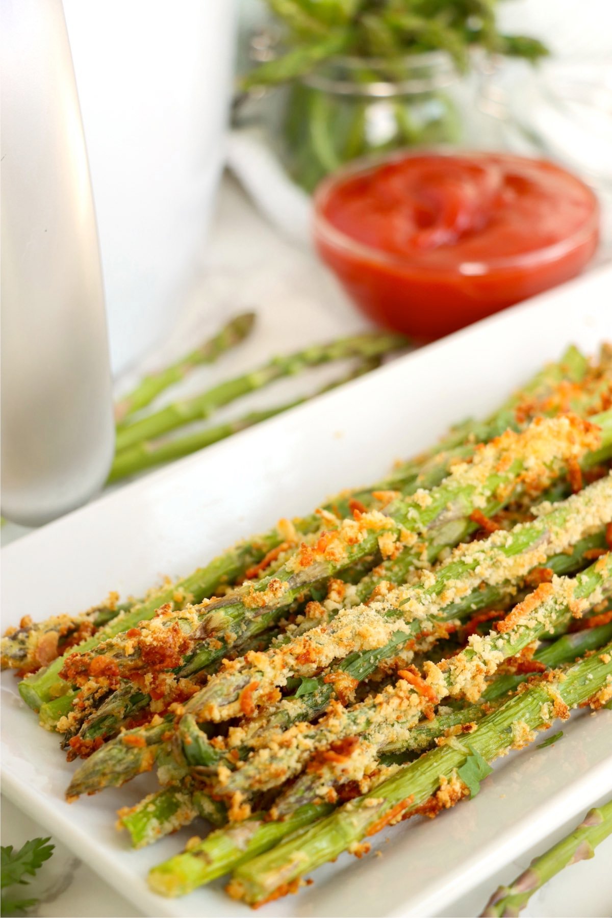 Side view of a tray of golden asparagus fries with a bowl of ketchup blurred in the background.