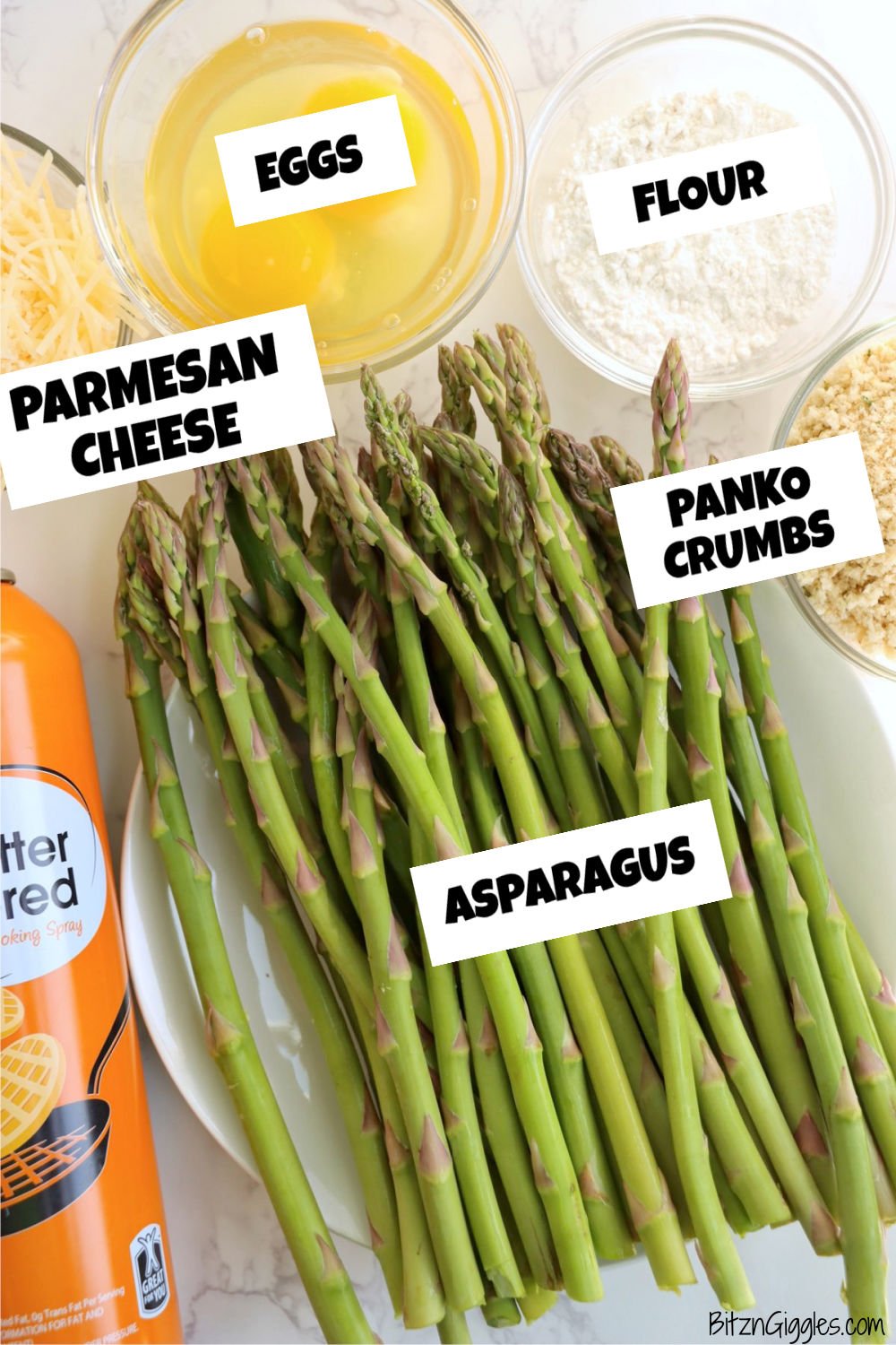 Fresh asparagus spears laid on a plate surrounded by small bowls of shredded cheese, beaten eggs, flour, and breadcrumbs, next to a can of nonstick cooking spray.