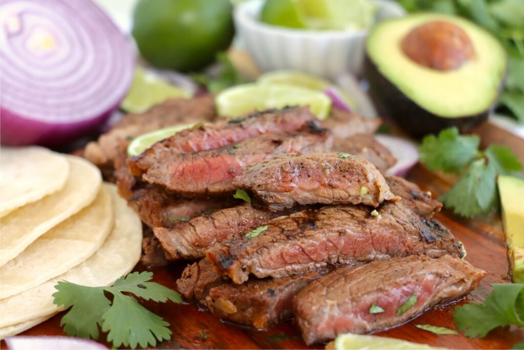 Sliced grilled steak with limes, avocado, and red onion, served on a wooden board.
