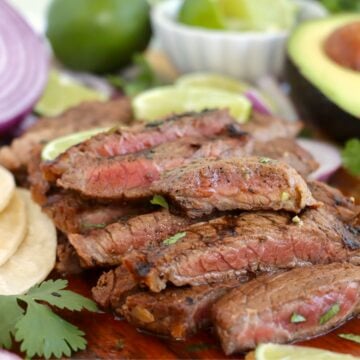 Sliced grilled steak with limes, avocado, and red onion, served on a wooden board.