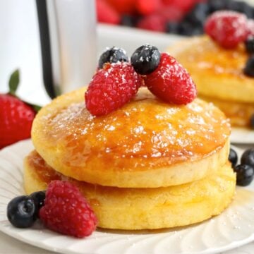 A horizontal view of golden pancakes with berries and syrup on a white plate, surrounded by scattered strawberries, raspberries, and blueberries.