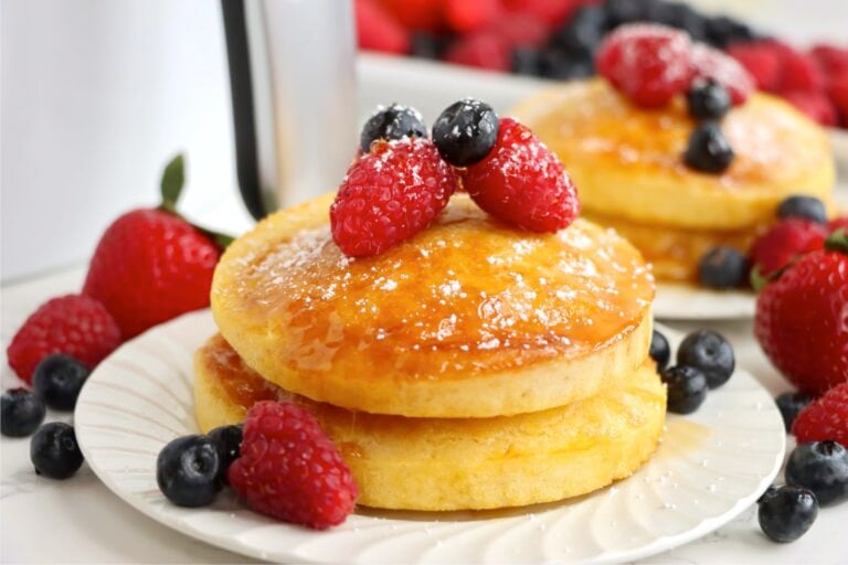 A horizontal view of golden pancakes with berries and syrup on a white plate, surrounded by scattered strawberries, raspberries, and blueberries.