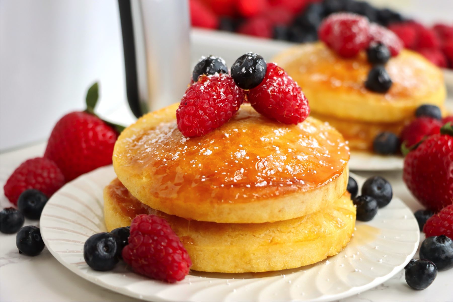 A horizontal view of golden pancakes with berries and syrup on a white plate, surrounded by scattered strawberries, raspberries, and blueberries.