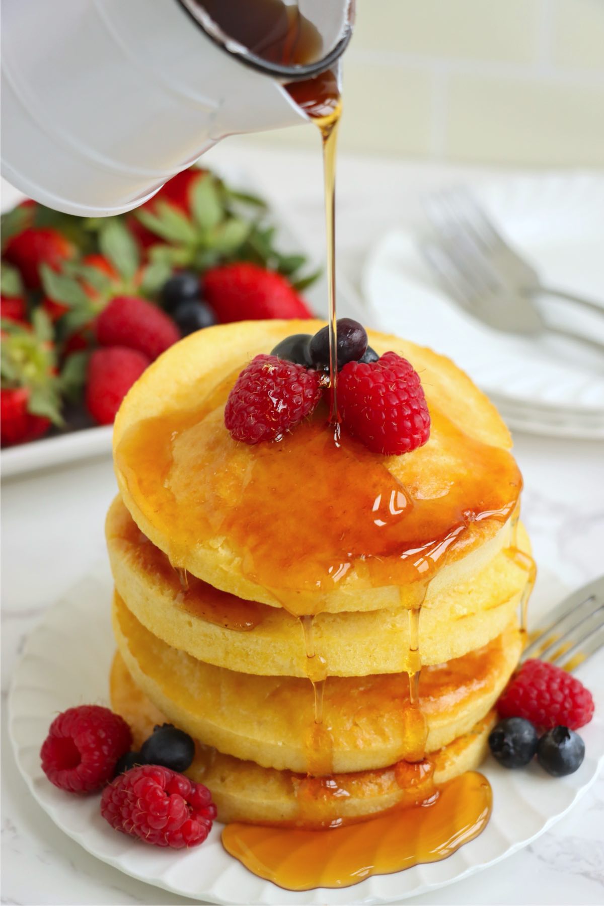A tall stack of golden pancakes topped with fresh raspberries and blueberries, as warm syrup is poured over them, cascading down the sides.