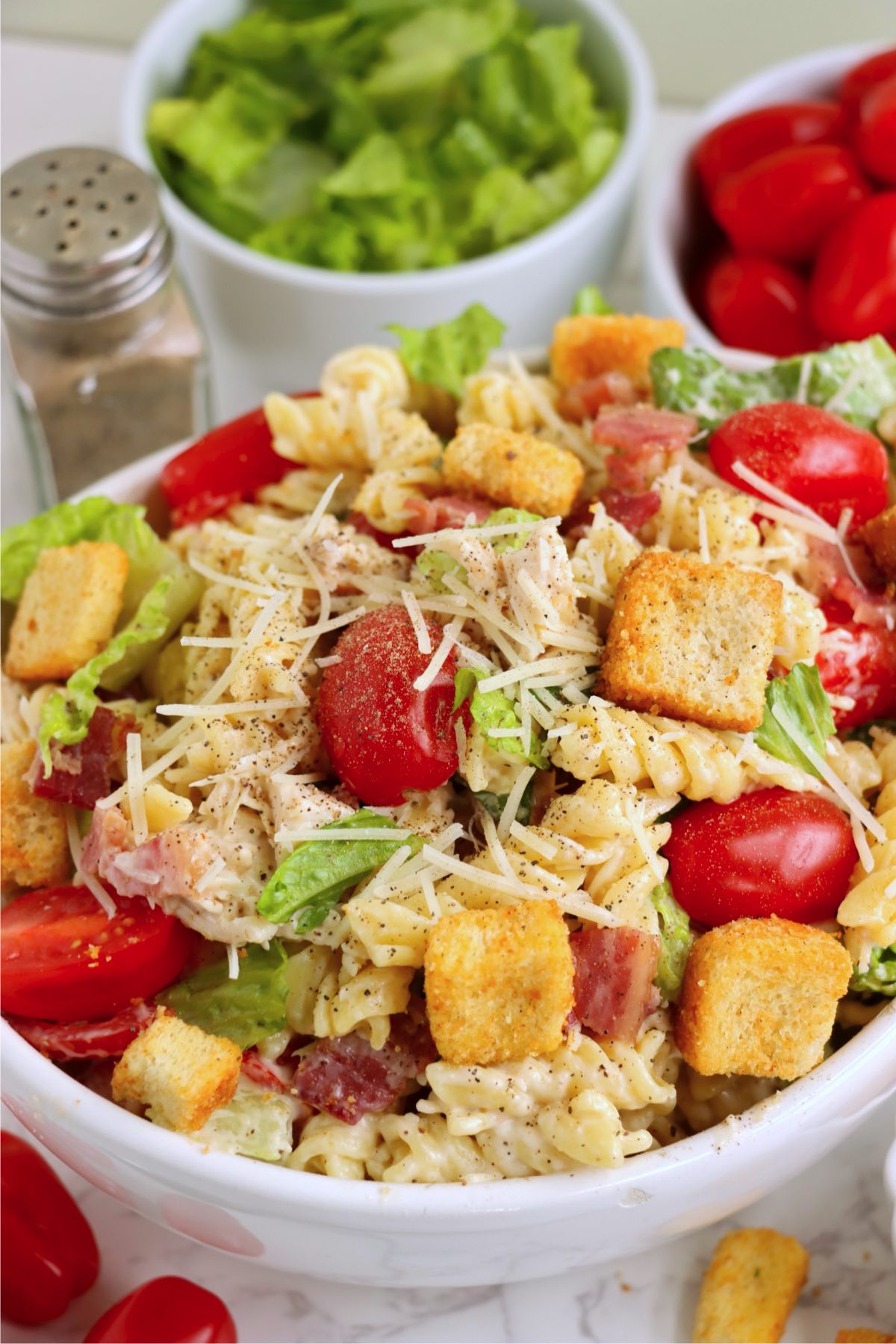Finished pasta salad in a bowl, surrounded by bowls of romaine and cherry tomatoes.