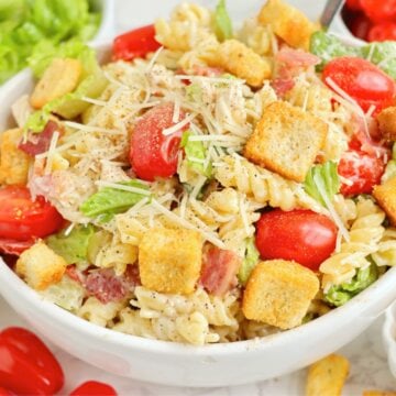 Pasta salad in a bowl, garnished with croutons and shredded parmesan, with tomatoes and lettuce in the background.