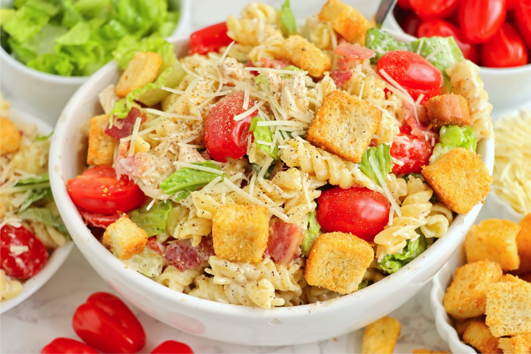 Pasta salad in a bowl, garnished with croutons and shredded parmesan, with tomatoes and lettuce in the background.