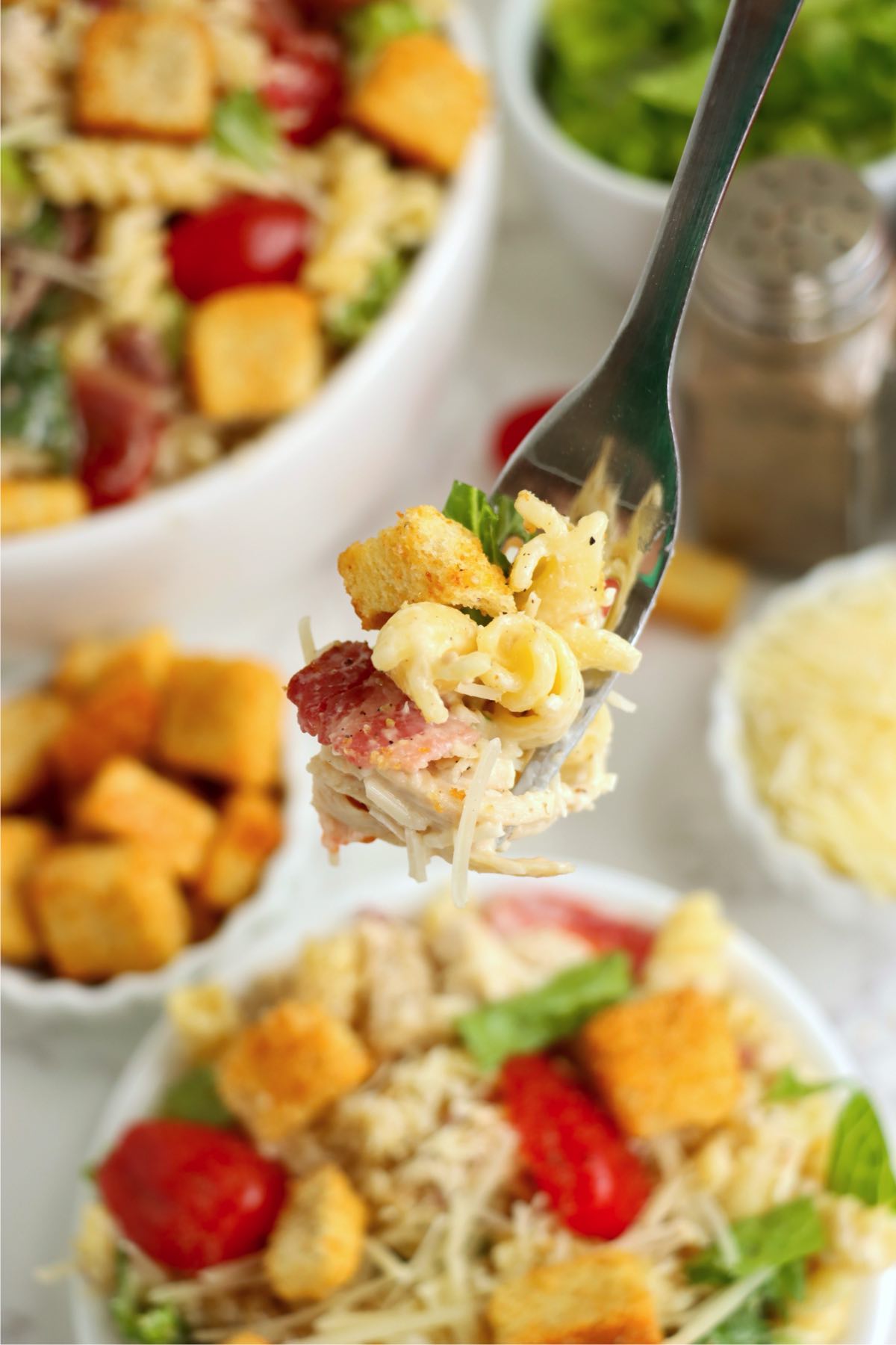 A forkful of chicken Caesar pasta salad, featuring rotini pasta, chicken, bacon, lettuce, parmesan, and a crouton, with the full salad bowl and ingredients blurred in the background.