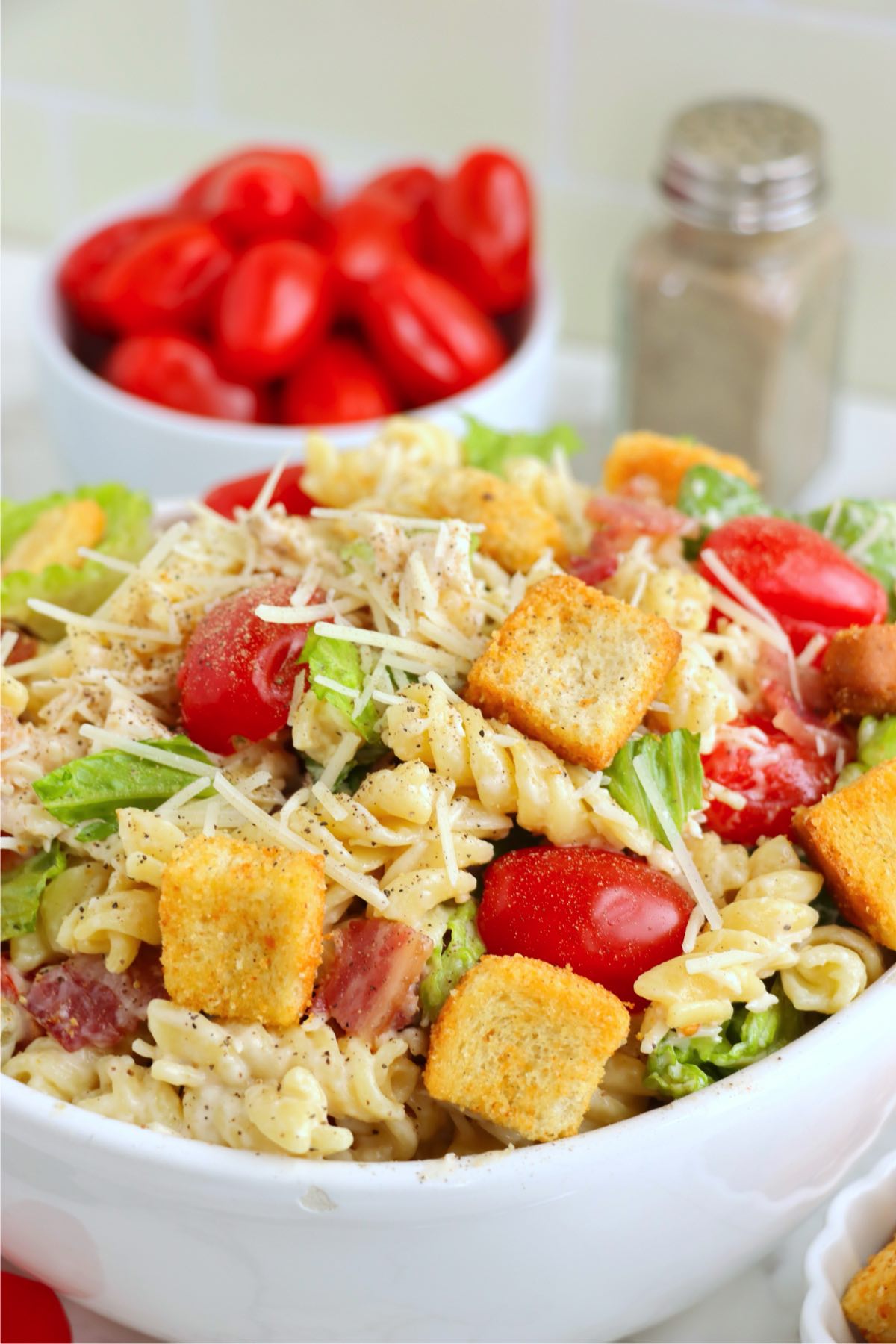 Close-up of a bowl of fully mixed chicken Caesar pasta salad topped with croutons and black pepper.