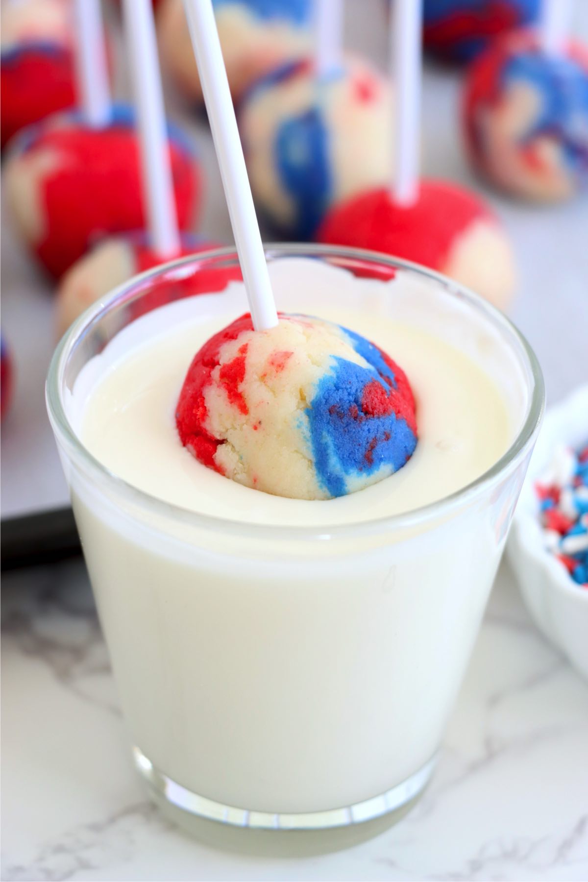 A tri-color cake pop on a stick being dipped into melted vanilla bark.