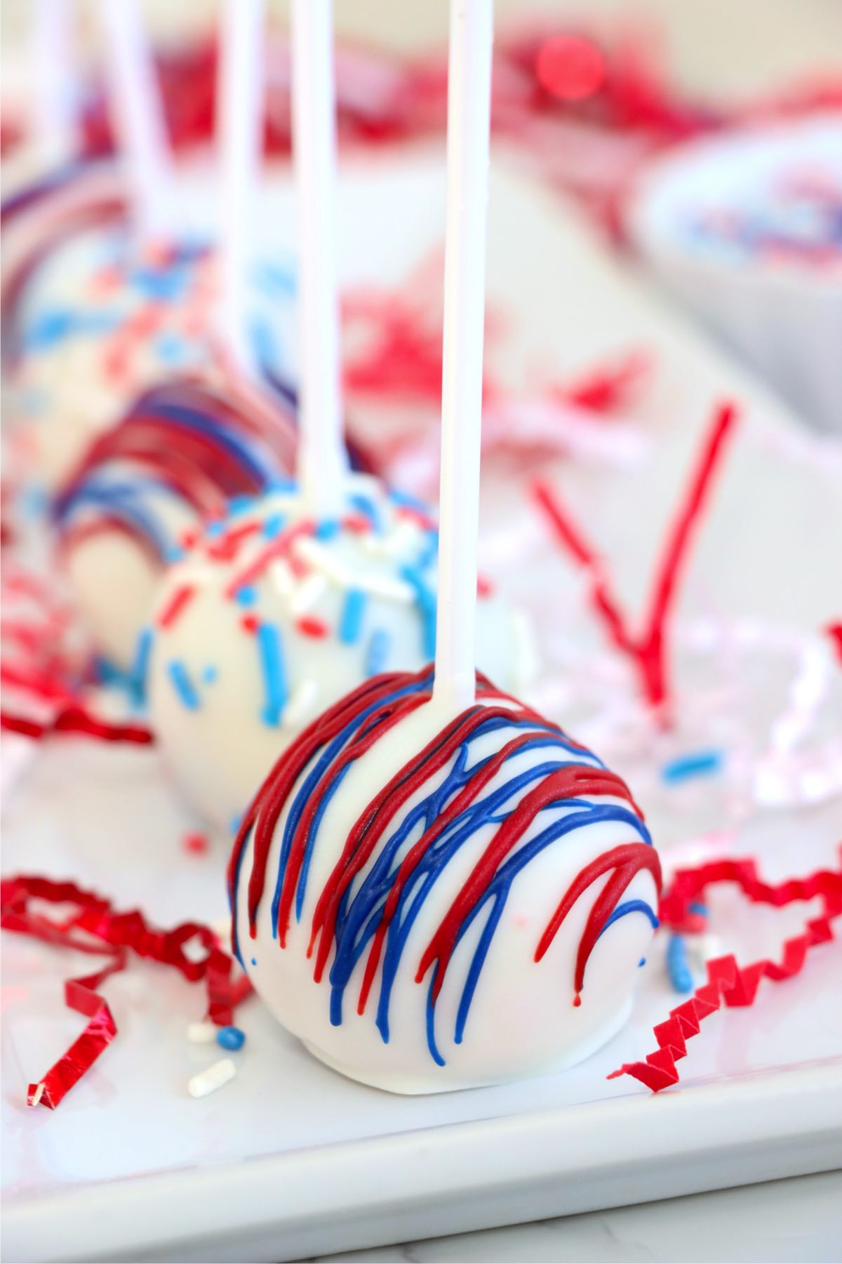 Frosted cake pops with patriotic drizzle and colorful sprinkles on a tray.