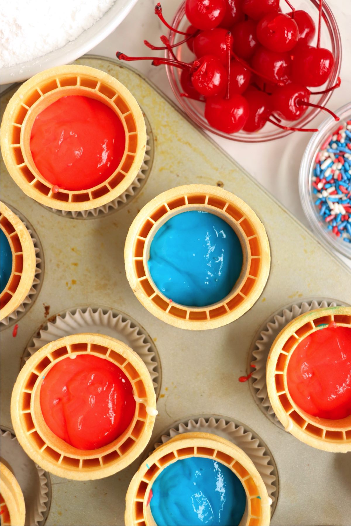 Ice cream cones in a muffin tin filled with red and blue cupcake batter, ready for baking.