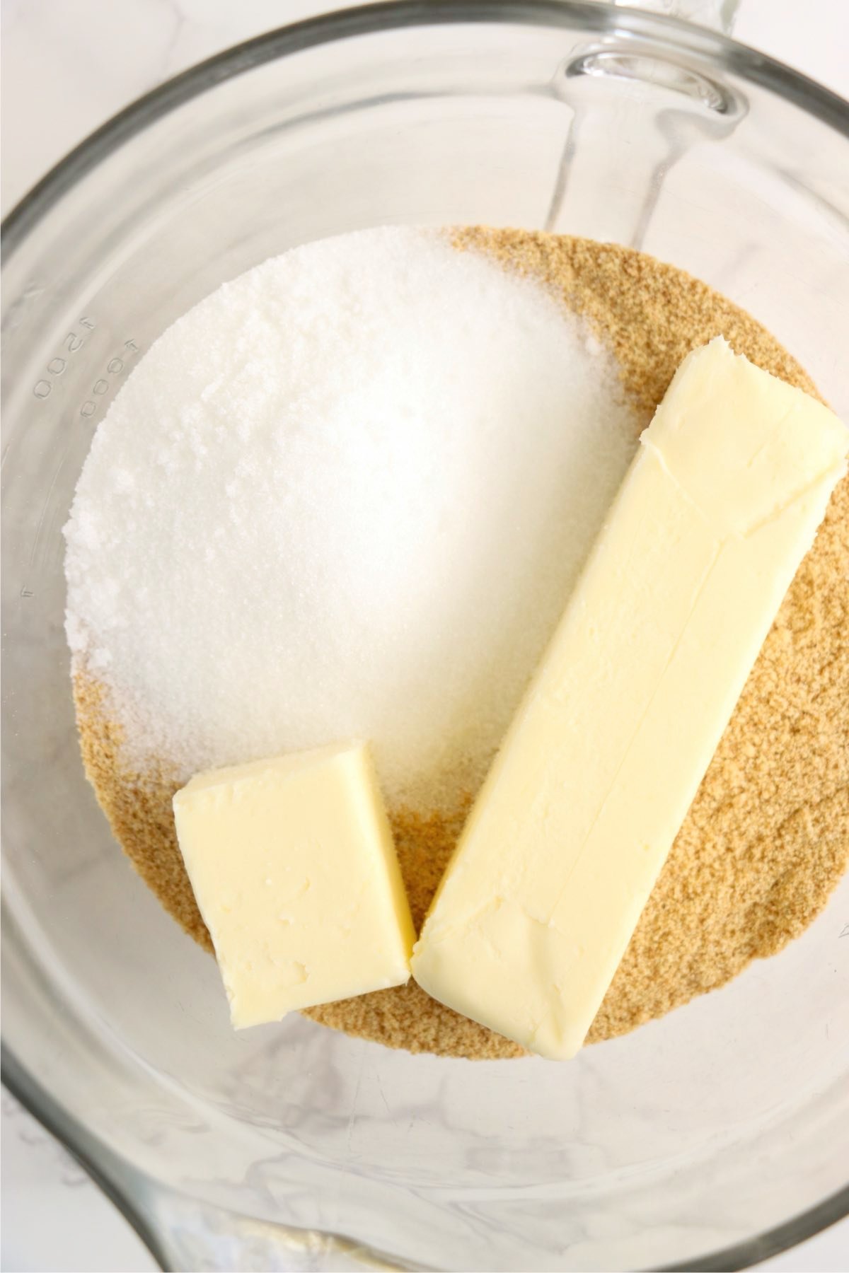 Graham cracker crumbs, sugar, and butter in a mixing bowl on a white surface.