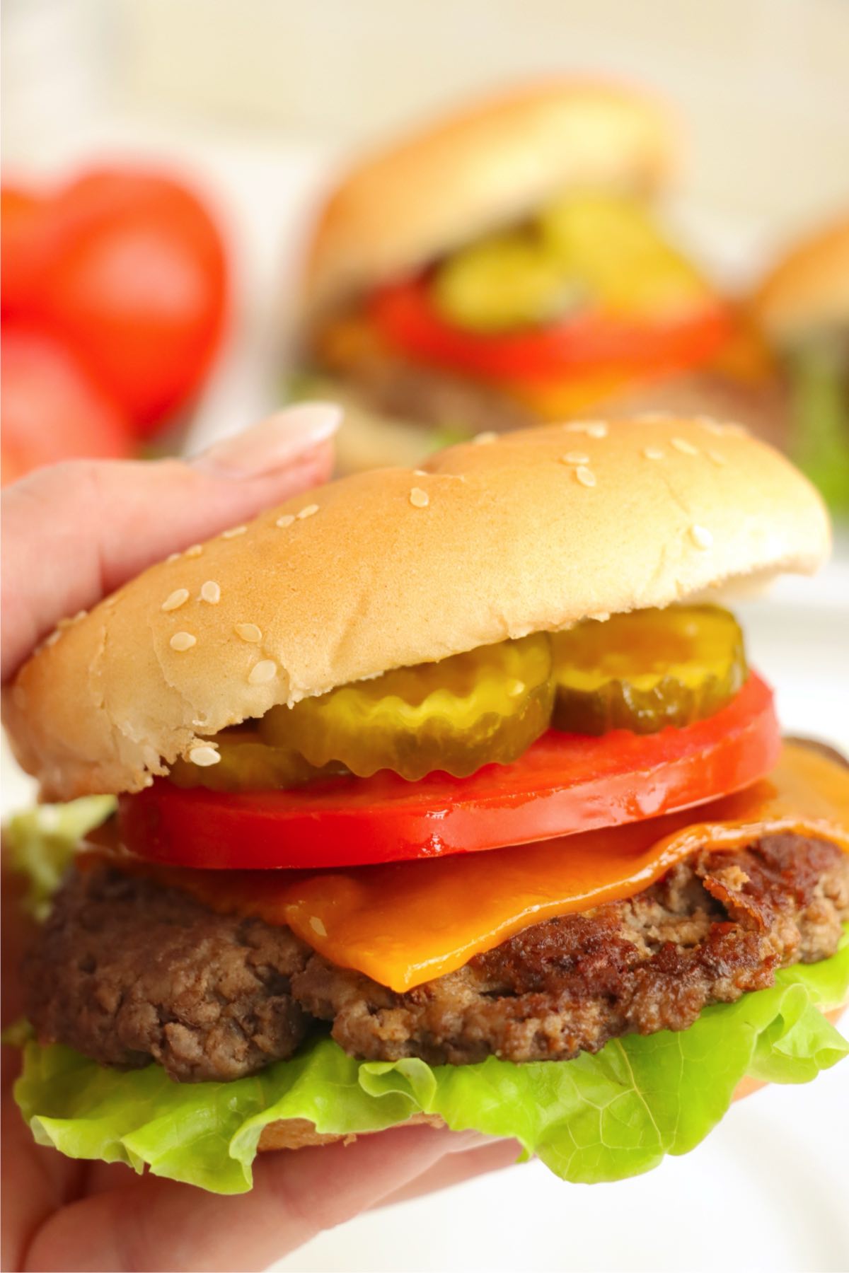 Hand holding a cheeseburger with lettuce, tomato, pickles, and cheddar cheese.
