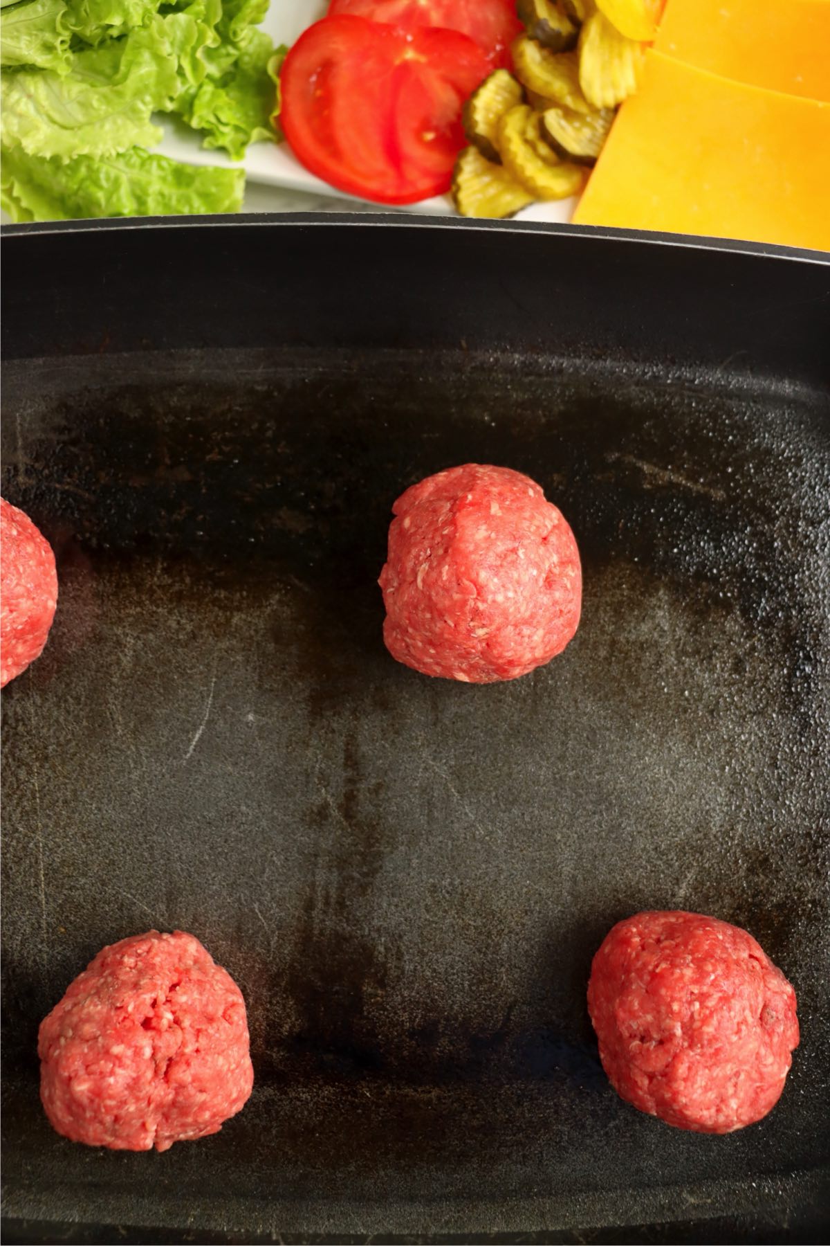 Four ground beef balls on a hot griddle, toppings in background.