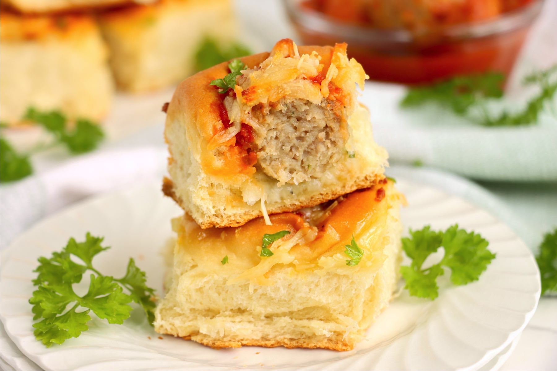 Stacked sliders on a white plate with fresh parsley garnish and a blurred background of more sliders.