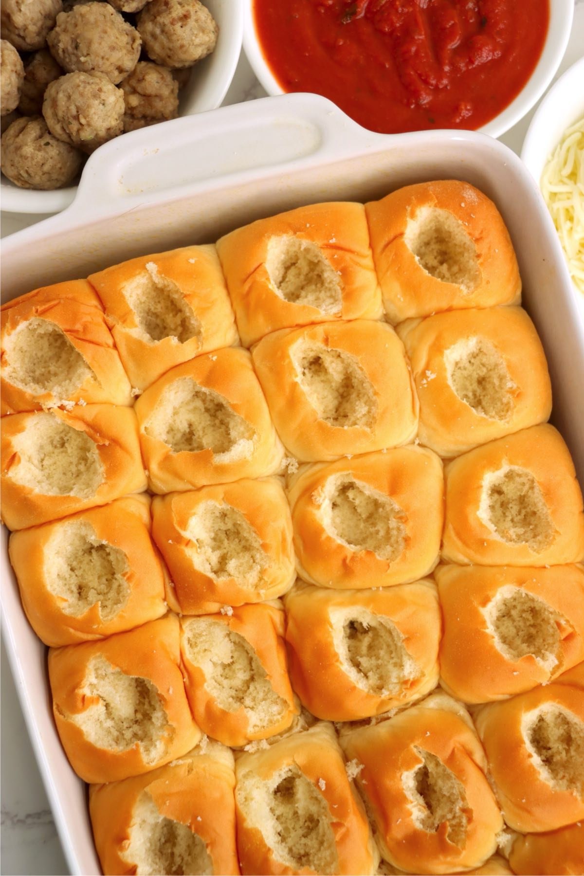 A baking dish of soft rolls with the centers carefully hollowed out, surrounded by bowls of meatballs, marinara, and shredded cheese.