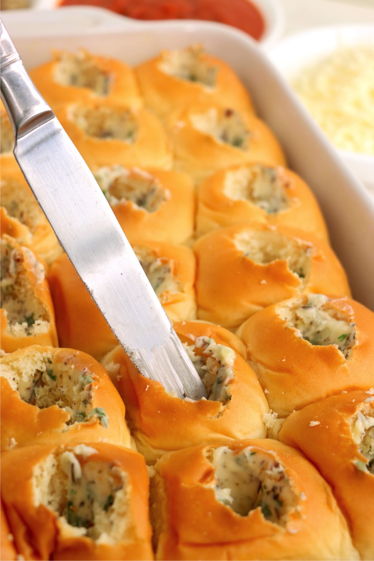 A butter knife spreading a herbed garlic butter mixture inside hollowed rolls in a baking dish.