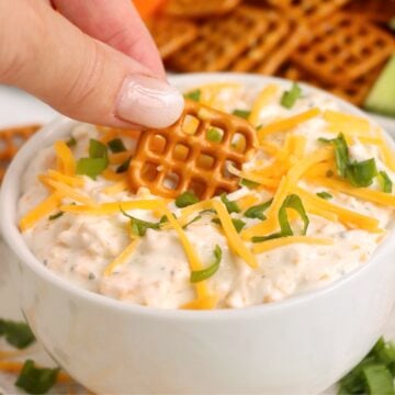 A pretzel bite being dipped into a bowl of beer dip garnished with cheese and green onions.