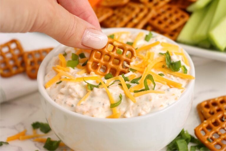 A pretzel bite being dipped into a bowl of beer dip garnished with cheese and green onions.