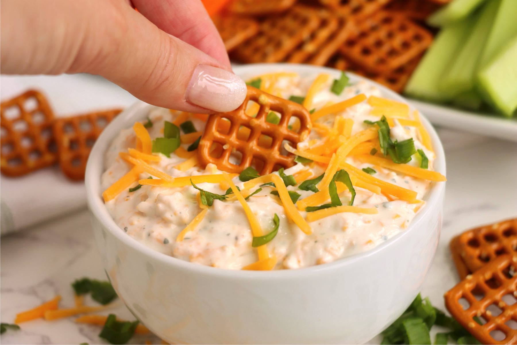 A pretzel bite being dipped into a bowl of beer dip garnished with cheese and green onions.