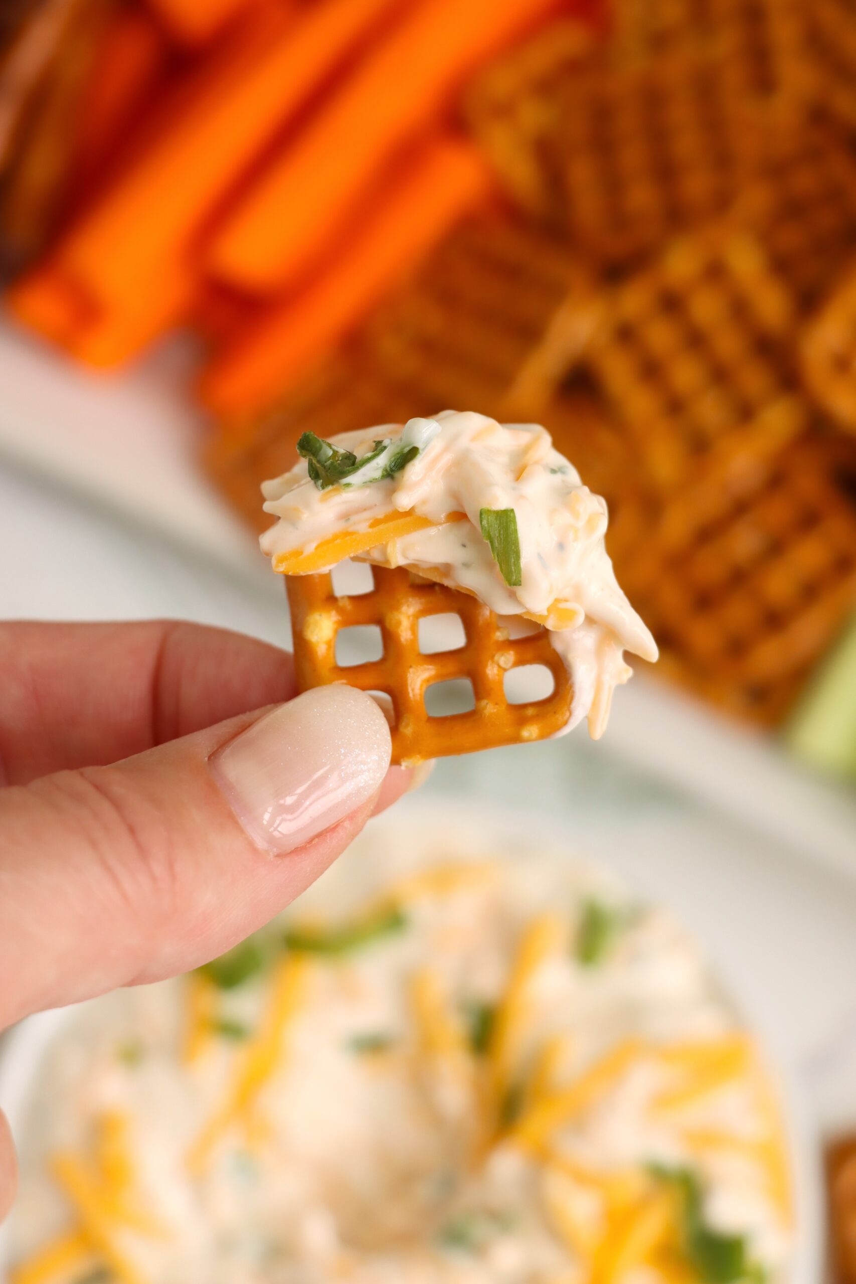 A hand holding a pretzel bite topped with ranch beer dip, cheddar, and green onion.
