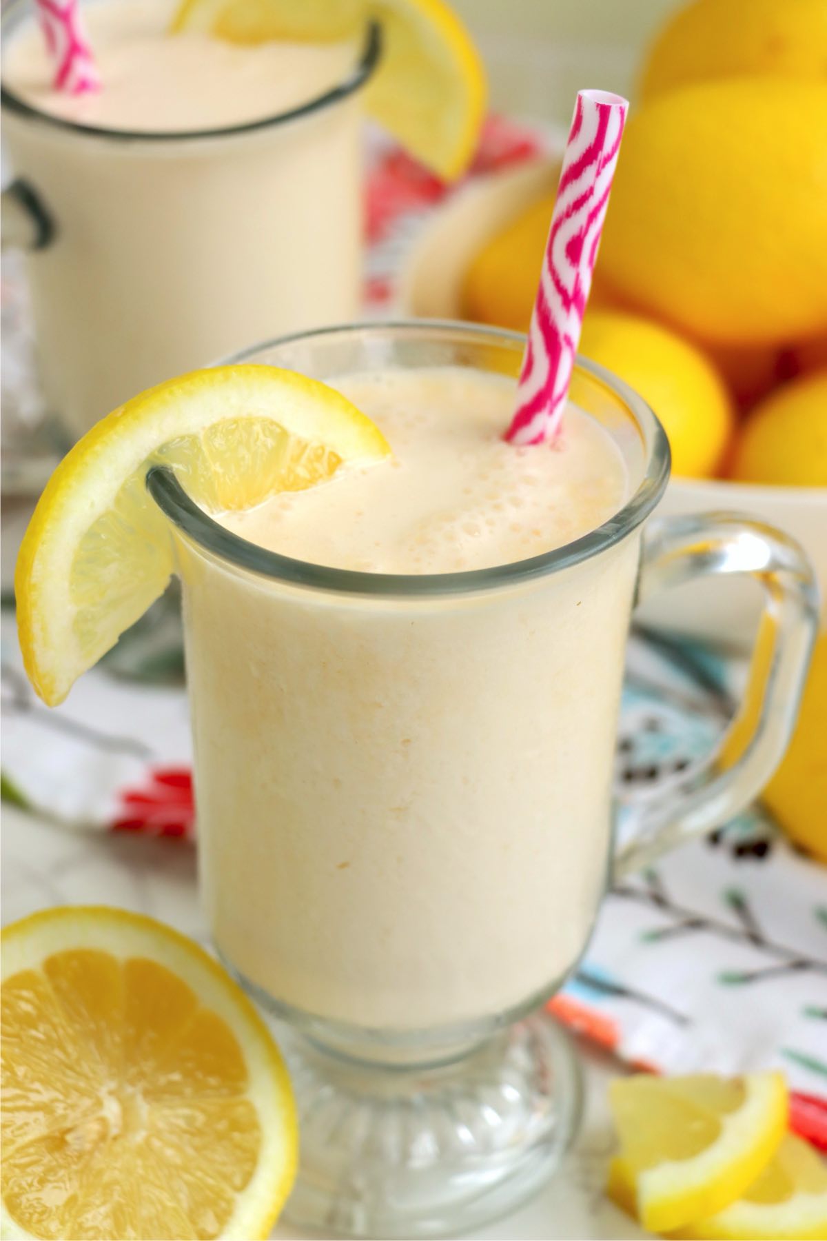 Two glass mugs of frosted lemonade on a colorful floral napkin, each garnished with a lemon slice and a pink-striped straw.